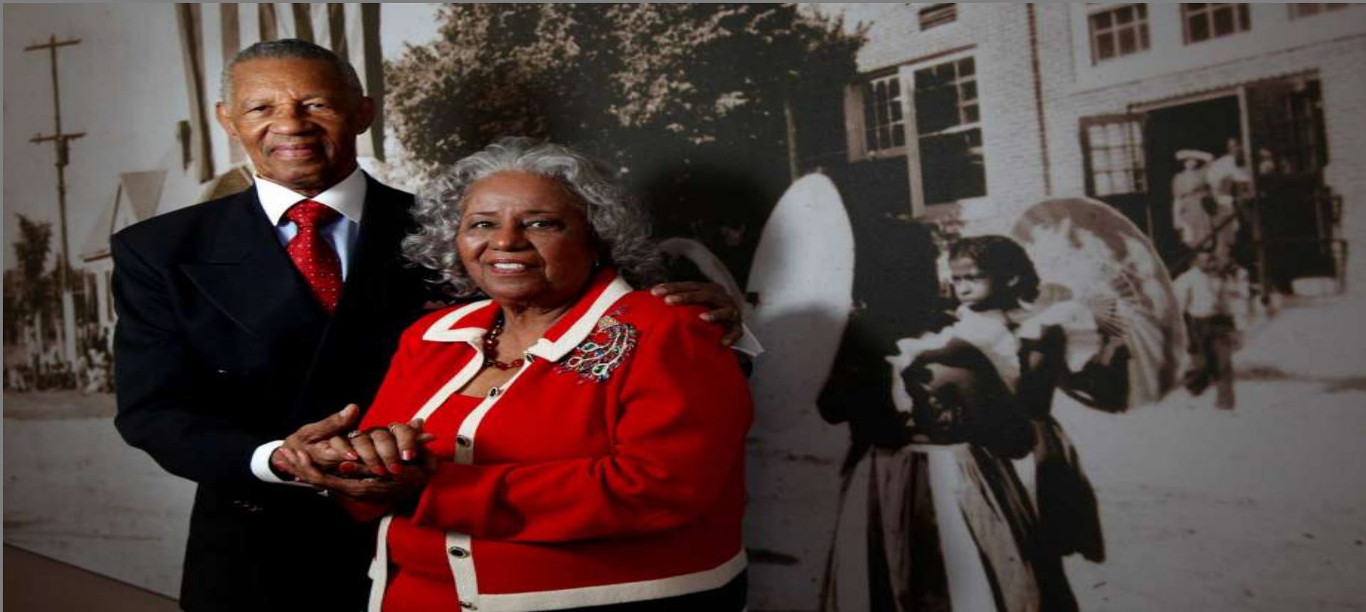 Elderly woman in red posing with historical photo background.